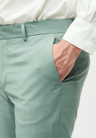Χέρι στην τσέπη ενός ανοιχτό πράσινου ραμμένου παντελονιού, που φοριέται με ένα λευκό μακρυμάνικο πουκάμισο με ορατό μανσέτι.