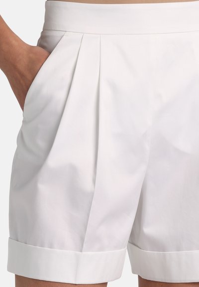 Shorts blancs taille haute, ajustés, avec des plis à l'avant et des ourlets retournés, portés avec la main gauche dans la poche, sur un fond neutre.