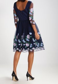 Mörkblå klänning med genomskinliga trekvartslånga ärmar, blommig broderi i pastellfärger, figursydd överdel, utsvängd kjol och dragkedja bak.