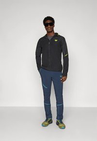 Sort let jakke med lynlås og stribet design, kombineret med marineblå bukser med blå accenter og robuste sko med grønne detaljer.