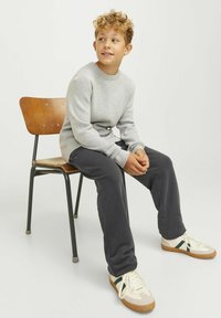 Grå tröja, mörka byxor och krämfärgade sneakers med gröna ränder. Sitter på en trästol mot en enfärgad bakgrund.