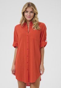 Robe chemise rouge en tissu léger, dotée d'un devant avec boutons, de manches roulées et d'une poche poitrine, avec un ourlet légèrement arrondi.