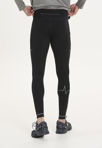 Zwarte leggings hebben een strakke pasvorm, ademende mesh accenten, een hartslagpatroon nabij de enkel en een ritszak aan de achterkant.