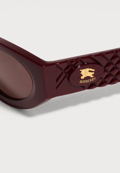 Gros plan sur une branche de lunettes de soleil Burberry bordeaux brillant, ornée d’un motif matelassé et d’un logo doré, sur fond blanc.