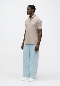 Beżowa koszulka polo z kołnierzykiem, krótkimi rękawami i logo. Połączona z jasnoniebieskimi spodniami typu wide-leg i białymi sneakersami. Gładkie tło.