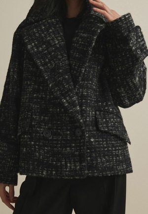 Woman wearing a black and gray plaid wool coat with wide lapels, button details on cuffs, and front flap pockets.