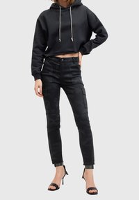 Femme portant un sweat à capuche court noir, un pantalon cargo camouflage noir et des sandales à talons hauts noires, debout avec une main sur la hanche.