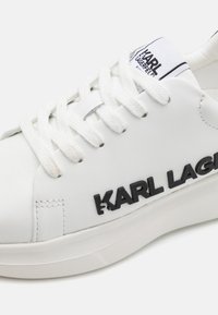 Bijela tenisica od teksturirane kože, s ravnim vezicama i crnim logotipom "KARL LAGERFELD". Ima podstavljeni ovratnik i teksturiranu gumu na potplatu.