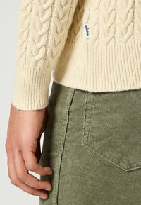 Beżowy sweter w splot kablowy z prążkowanym dołem, zestawiony z oliwkowymi spodniami z szerokiego prążka o fakturowanej tkaninie i szczegółowych szwach.