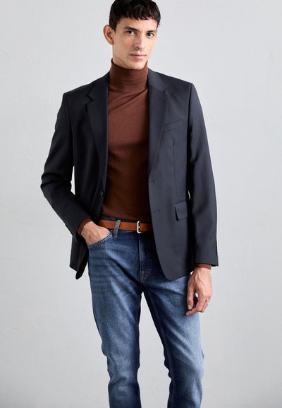 Blazer preto sobre um suéter de gola alta castanho, combinado com jeans de ganga azuis e um cinto castanho. O blazer possui um único bolso no peito e um corte ajustado.