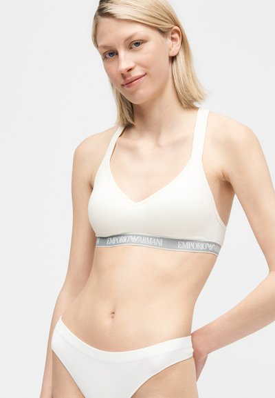 Νεαρή γυναίκα με κοντά ξανθά μαλλιά που φοράει λευκό αθλητικό σουτιέν Emporio Armani και ταιριαστά λευκά εσώρουχα σε ανοιχτό γκρι φόντο.