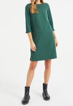 Frau, die ein knieumspielendes, dunkelgrünes Kleid mit dreiviertellangen Ärmeln und schwarze, klobige Stiefeletten trägt, steht vor weißem Hintergrund.