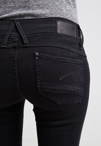 Black denim jeans with a fitted design, featuring a leather patch at the waist, back pockets with a subtle stitch detail, and a smooth texture.