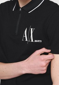 Personne portant un polo noir avec des bordures blanches et un logo blanc A|X Beats sur la poitrine, ajustant la manche de la chemise avec la main.