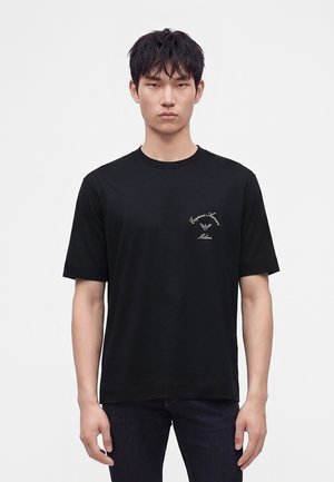 Jeune homme portant un t-shirt noir à manches courtes avec un petit logo blanc Emporio Armani sur la poitrine gauche, debout devant un fond uni.