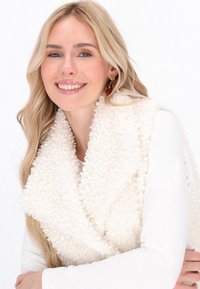 Blonde vrouw die glimlacht, met een wit, krullend vest over een lange mouw witte top, met hangende oorbellen tegen een witte achtergrond.