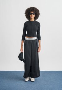 Μαύρο μακρυμάνικο πλεκτό τοπ με κουμπιά και λευκό λογότυπο Nike, σε συνδυασμό με άνετο μαύρο παντελόνι, κρατώντας μαύρο καπέλο και φορώντας σκουρόχρωμα γυαλιά ηλίου.