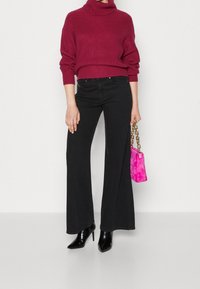 Nainen, jolla on burgundinvärinen poolopaita, mustat leveälahkeiset housut ja mustat korkokengät, pitää kädessään pientä pinkkiä pörr�öistä käsilaukkua, jossa on kultaketju.