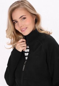 Mustat fleece-takki, jossa on korkea kaulus ja vetoketjullinen etuosa, jossa on hillitty logo. Takki on päällä valkoisen ja mustan raidallisen paidan.