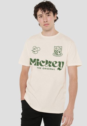 Young man with curly hair wearing a beige T-shirt with green "Mickey The Original" text and cartoon-style graphics, paired with black pants.