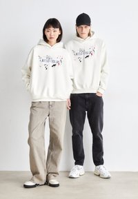 Des sweatshirts à capuche blancs avec un design éclaboussé coloré et un texte sur le devant, associés à des jeans en denim clairs et foncés. Des chaussures décontractées complètent le look.