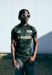 Zaļš kamuflāžas Adidas krekls ar dzelteniem akcentiem un logotipu, apvienots ar pelēkiem džinsu šortiem. Valkā baltas saulesbrilles un melnu kaklarotu.