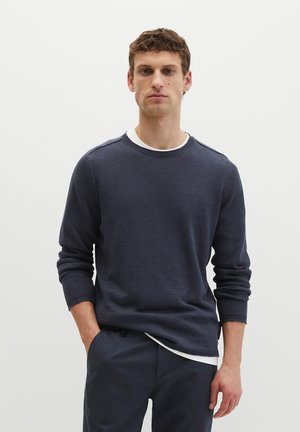 Młody mężczyzna stoi na jednolitym tle, ubrany w ciemnoniebieski sweter z okrągłym dekoltem na białą koszulkę oraz ciemnoniebieskie spodnie, jedną ręką w kieszeni.