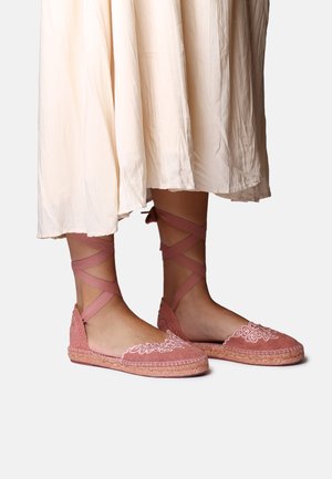 Jalat, joissa on vaaleanpunaiset espadrillolenkkarit kukkakirjailulla ja nilkan ympäri solmittavilla nauhoilla, yhdistettynä virtaavaan vanhan valkoiseen hameeseen.