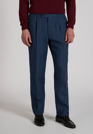 Uomo con pantaloni blu navy su misura, mocassini neri e maglia a maniche lunghe bordeaux, in piedi davanti a uno sfondo neutro.