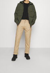 Olivgrön zip-jacka med två framfickor, svart t-shirt, beige rak passformbyxor och svarta skor. Slät tyg med avslappnad passform.