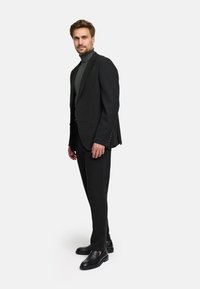 Traje negro a medida con pantalones a juego, llevado sobre un jersey de cuello alto verde oscuro. Los zapatos son negros, de punta redonda con un ligero detalle decorativo.