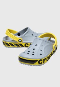 Szare klapki Crocs z żółtymi akcentami, z otworami wentylacyjnymi, pasem z tyłu oraz wzorzystym czarnym paskiem wzdłuż podeszwy.