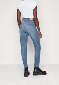 Modré džínové džíny s vysokým pasem a zúženým střihem, s koženou logovou nášivkou na zadní straně a dvěma zadními kapsami. Nošeny s černými kotníkovými botami.