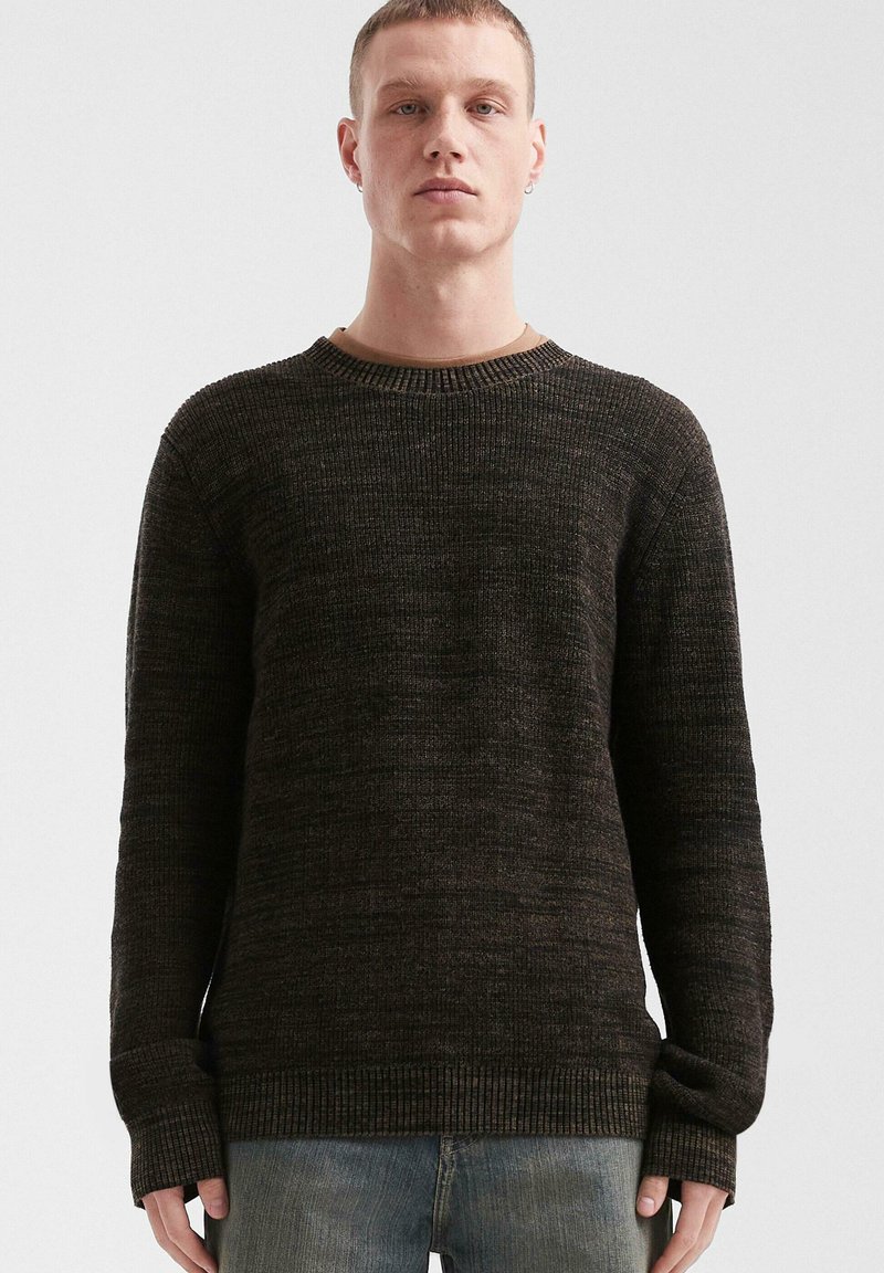 Junger Mann mit kurzen Haaren, der einen dunkelbraunen Strickpullover und verwaschene blaue Jeans trägt, steht vor einem schlichten, hellen Hintergrund.