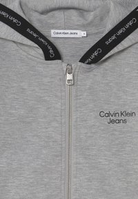 Szara bluza z kapturem na zamek błyskawiczny o gładkiej teksturze, z metalowym zamkiem i czarnym sznurkiem do ściągania. Na przodzie znajduje się logo "Calvin Klein Jeans".