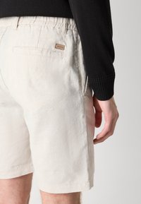 Beige vászon rövidnadrág elasztikus derékkal és hátsó zsebbel, egy kis barna márkajelzéssel. Az anyaga puha és enyhén gyűrött.