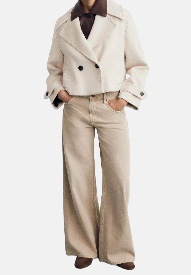 Personne portant un manteau court beige à double boutonnage par-dessus une chemise marron foncé, un pantalon beige à jambes larges et des chaussures marron, les mains dans les poches.