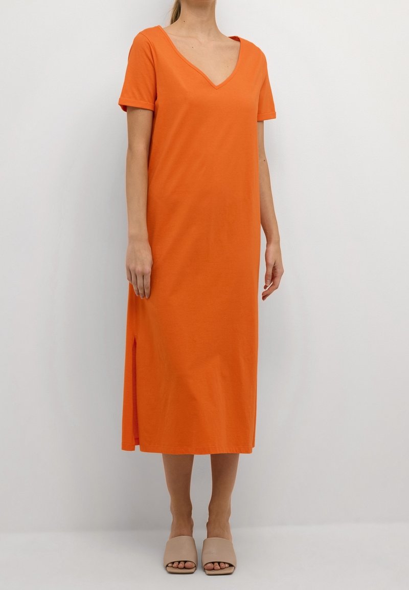 Femme portant une robe midi orange à manches courtes en V avec fentes latérales et des chaussures beige ouvertes à enfiler, debout devant un fond uni.