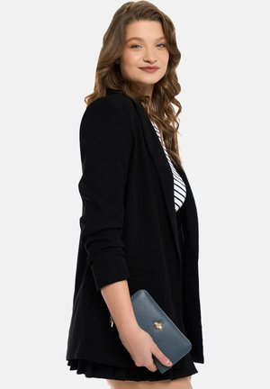 Schwarze Blazer über einem gestreiften weißen Oberteil, hält eine strukturierte blaue Geldbörse mit einem goldenen Bienen-Akzent. Der Hintergrund ist rein weiß.