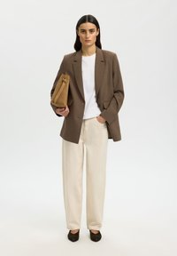 Žena stojící, oblečená v hnědém blejzru, bílém tričku, krémových kalhotách a černých botách, držící velkou hnědou semišovou kabelku na bílém pozadí.