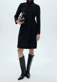 Abrigo de lana negro con un alto cuello, diseño de doble botonadura y dos bolsillos laterales, combinado con botas de cuero negras hasta la rodilla.