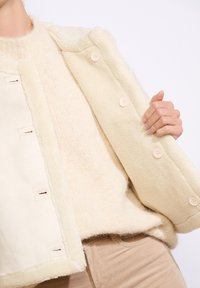 Crèmekleurige fleece vest met een zachte textuur, voorzien van vijf grote beige knopen en een pluche kraag, gelaagd over een lichte gebreide trui.