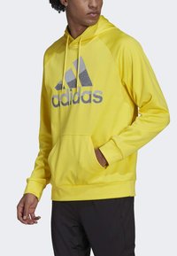 Felpa gialla con cappuccio realizzata in tessuto morbido, con un grande logo Adidas grigio sulla parte anteriore, tasca a marsupio e polsini a costine.
