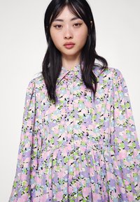 Vestido floral com uma base lavanda e detalhes em rosa, verde e preto. Gola abotoada e mangas longas com detalhes franzidos.