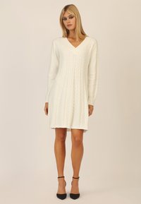 Witte gebreide jurk met een V-hals, lange mouwen en een kabelbreipatroon. Afgewerkt met een uitlopende zoom en gedragen met zwarte stilettos.