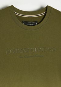 T-shirt w kolorze oliwkowym z okrągłym dekoltem, z haftowanym napisem "DIVERSE HERITAGE" oraz "True Original Clothing" w kontrastowej nici.