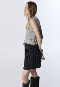 Femme de profil portant un gilet sans manches gris, une jupe plissée grise avec cordon rouge, et des bottes noires jusqu'aux genoux, se tenant contre un fond blanc.