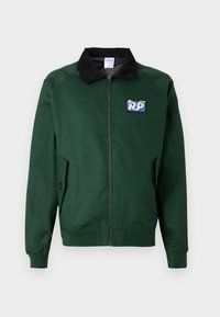 Zelena bomber jakna s crnim kordura ovratnikom, prednjim zatvaračem, bočnim džepovima i plavom naljepnicom s natpisom "R.P." na lijevoj strani prsa.