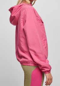 Osoba ubrana w jasnoróżową kurtkę z kapturem i elastycznymi mankietami, w połączeniu z beżowymi spodniami z dużą różową łatą na udzie.