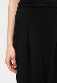 Κοντινή λήψη του χεριού ενός ατόμου που ακουμπάει δίπλα σε μαύρο πλισέ ψηλόμεσο παντελόνι και μαύρο τοπ σε μονόχρωμο φόντο.